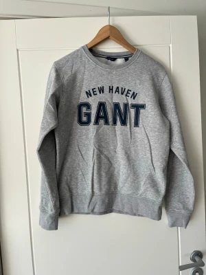 Grå sweatshirt från GANT - Snygg grå sweatshirt från GANT med tryck 'NEW HAVEN GANT' i mörkblått på bröstet. Tröjan har rund halsringning med ribbad kant och är tillverkad i mjukt bomullsmaterial. Perfekt för en avslappnad och cool look.priset kan diskuteras 