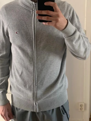 Grå ziptröja från Tommy Hilfiger - Snygg grå ziptröja från Tommy Hilfiger i premium bomull. Tröjan har hel dragkedja, ribbade muddar och diskret logga på bröstet. Kragen har coola ränder i rött, vitt och svart på insidan. Perfekt till ett avslappnat men stilrent uttryck.