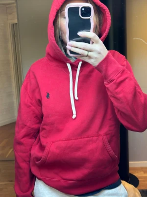 Röd hoodie från Polo Ralph Lauren - Röd hoodie från Polo Ralph Lauren med vit dragsko i huvan och klassisk broderad logga på bröstet. Mjuk bomullskänsla och känguruficka framtill. Perfekt för en avslappnad och stilren look. Sitter som en S/M