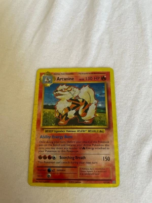 Arcanine Pokémonkort Holo - Säljer ett holografiskt Arcanine Pokémonkort med gul ram och färgglad illustration. Kortet har 130 HP och visar Arcanine i en naturmiljö. Text och detaljer på engelska, baksidan har klassisk blå Pokémon-design med Pokéboll.