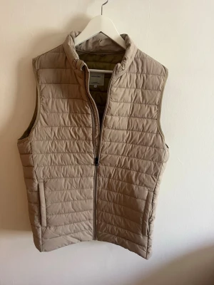 Beige dunväst från jack&jones - Stilren beige dunväst från Jack&Jones med quiltad design och hög krage, storlek medium. Västen har dragkedja framtill och två sidofickor. Perfekt för lager-på-lager och enkel att matcha med olika outfits.