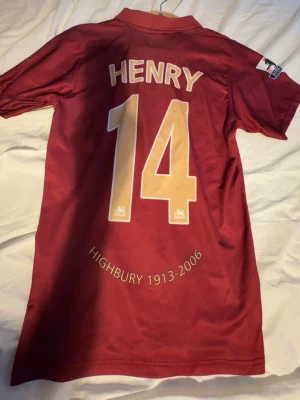Arsenal Highbury tröja Henry 14 - Säljer en vinröd Arsenal Highbury 1913-2006 matchtröja från Nike med Henry 14 på ryggen. Tröjan har klubbmärke, O2 sponsortryck och Premier League-märke på ärmen. Klassisk pikékrage och korta ärmar. Perfekt för fotbollsfans!