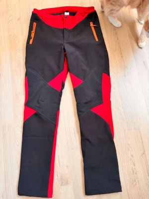 Svarta och röda softshellbyxor - Snygga softshellbyxor i svart och rött med orange dragkedjor. Byxorna är fleecefodrade och därför mycket varma! Byxorna har förstärkta partier på knäna och stretchigt material för extra rörlighet. De är knappt använda och därav i nyskick! Finns någon enstaka fläck som går bort i tvätten. Det står storlek L på lappen men byxorna är väldigt små i storlek och har varit snäppet för stora för mig som annars har S eller 36. Skulle uppskatta storleken till 38.