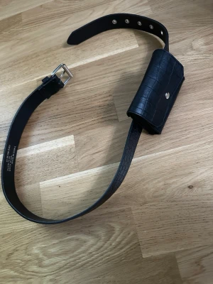 Svart bälte med väska i krokopräglat skinn - Snyggt svart bälte med liten väska i krokopräglat skinnimitation. Perfekt för att bära småsaker och ger en trendig touch till din outfit. Väskan har silverfärgad knappstängning och bältet har klassiskt spänne.