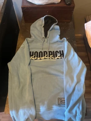 Ljusblå HOODRICH hoodie med tryck - Säljer en ljusblå hoodie från HOODRICH med stor svart och vit logga på bröstet och en patch nertill. Hoodien har huva med dragsko, magficka och mjukt material på insidan. Perfekt för dig som gillar streetwear och vill ha en chill look.