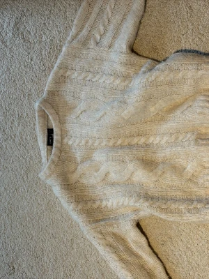 Beige kabelstickad tröja  - Kabelstickad tröja i mjuk, vit ullmix.Tröjan har ett klassiskt flätmönster, rund halsringning och långa ärmar. Perfekt för lager på lager och en stilren look.