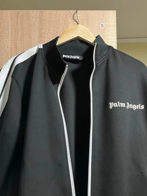 Palm Angels svart track jacket - Palm Angels svart track jacket med vita detaljer och logga på bröstet. Jackan har ståkrage, dragkedja framtill och vita ränder längs ärmarna. Tillverkad i 80% bomull och 20% polyester för en skön och sportig känsla. Använt ett par gånger säljer nu för den inte är min still 
