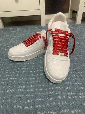 Nike Air Force 1 x Supreme vita sneakers - Fräscha vita Nike Air Force 1 sneakers med röda Supreme-skosnören. Klassisk låg modell i läder med perforerad tåbox och tydlig Nike Swoosh på sidan. Riktigt clean look med en twist tack vare de röda snörena med Supreme-logga.