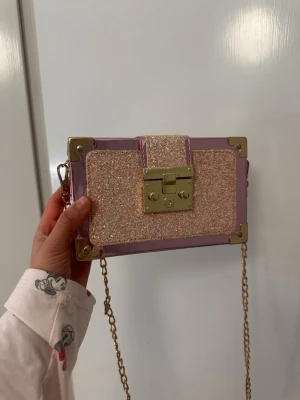 Glittrig rosa kuvertväska med kedja - Säljer en superfin kuvertväska i boxmodell med glittrig rosa och beige framsida, guldfärgade metalldetaljer och kedjerem. Väskan har ett dekorativt lås framtill och hårda hörn i metall. Perfekt för dig som vill sticka ut med accessoarer!