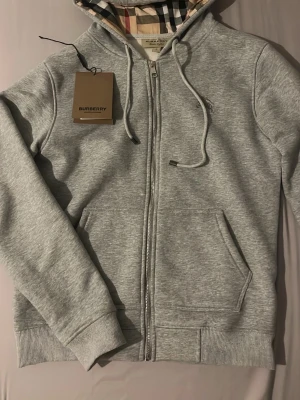 Grå zip hoodie från Burberry - Snygg grå zip hoodie från Burberry med klassiskt rutigt foder i huvan och dragsko. Mjuk bomullsblandning, ribbade muddar och fickor framtill. Perfekt för en chill och stilren look.