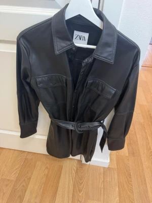 Svart skinnkappa från Zara - Snygg svart kappa i skinnimitation från Zara med bälte i midjan och stora bröstfickor. Kappan har klassisk krage, knäppning framtill och långa ärmar. Perfekt för dig som vill ha en trendig och edgy look. Stl xs