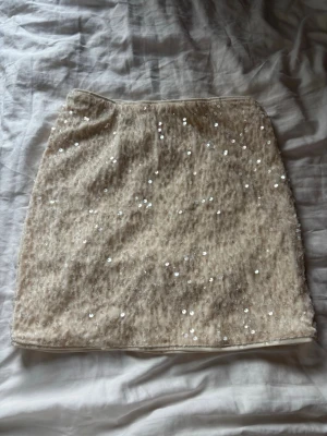 Beige paljettkjol i kort modell - Trendig beige kortkjol täckt med glittriga paljetter. Kjolen har en rak passform och är perfekt för dig som vill sticka ut lite extra. Materialet känns mjukt och skimrande, vilket ger en lyxig känsla. Storlek 134/140