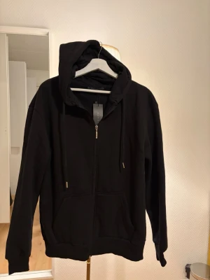 Svart zip hoodie med huva - Snygg svart hoodie med dragkedja framtill och stor huva. Hoodien har dragsko i huvan, två fickor fram och ribbade muddar vid ärmslut och nederkant. Perfekt för en chill och avslappnad stil.