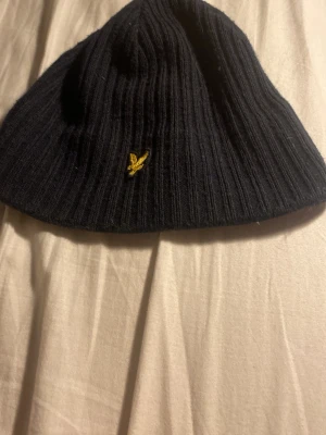 Svart ribbad mössa från Lyle & Scott - Säljer en svart ribbstickad mössa från Lyle & Scott med den klassiska gula örnloggan broderad framtill. Mössan är mjuk och stretchig, perfekt för kalla dagar och enkel att matcha med olika outfits.