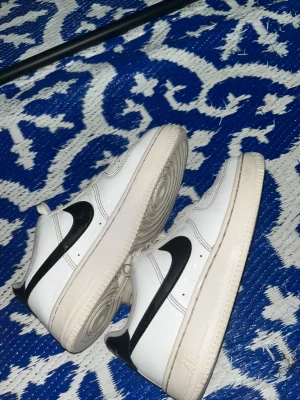 Nike Air Force 1 vita och svarta sneakers som är knappt använda, använda ett par gånger  - Knappt använda, använda ett par gånger. Klassiska Nike Air Force 1 sneakers i vitt läder med svart swoosh på sidorna. Skorna har perforerad tå för ventilation, vita skosnören och en tjock vit sula med Air-dämpning. Perfekt för dig som gillar tidlös streetstil.