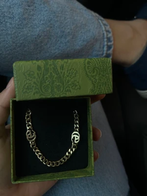 Guldigt kedjearmband med logodetaljer - Snyggt armband i guldfärgad metall med kraftig kedja och två dekorativa logodetaljer. Kommer i en grön mönstrad ask, perfekt för dig som gillar stilrena accessoarer med en twist. Passar till många olika looks och är lätt att matcha.