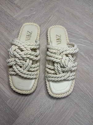 Krämvita sandaler med repdetaljer Zara - Unika sandaler från Zara i krämvitt med chunky repdetaljer över foten. Sandalerna har fyrkantig tå och öppen häl, med flätad design i textil som ger en cool och trendig look. Perfekta för dig som vill sticka ut med din stil.