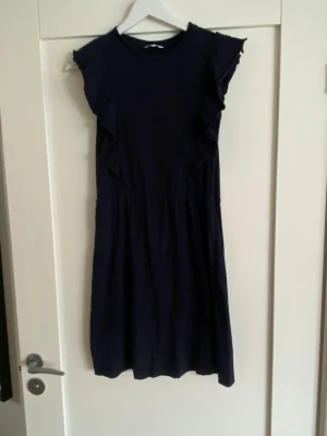 Marinblå klänning med volangärmar - En enkel och stilren marinblå klänning med volangdetaljer längs ärmarna. Klänningen har rund halsringning och är tillverkad i mjuk bomull, vilket gör den bekväm att bära. Passformen är normal och längden är kort, perfekt för varmare dagar.