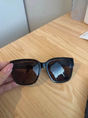 Bruna/Svarta solglasögon från Balenciaga - Stilrena svart-bruna solglasögon från Balenciaga med bred båge och fyrkantig form. Glasen är mörka och bågarna har en diskret logga på utsidan. Perfekta för att lyfta vilken outfit som helst.  