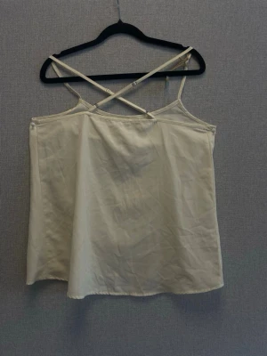 Beige linnetopp med korsade band - Stilren beige linnetopp med tunna axelband som korsas i ryggen. Toppen har en enkel och rak passform, perfekt för varma dagar. Materialet känns lätt och svalt, vilket gör den skön att bära. Snygg att matcha med jeans eller kjol.
