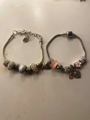 Armband med berlocker - Säljer två snygga silvriga armband med flera olika berlocker, bland annat hjärtan, blommor och pärlor i rosa och silver. Armbanden har ormkedja och detaljer i metall samt glittrande och mönstrade pärlor. Perfekt för dig som gillar personliga accessoarer. 1 armband 30kr, båda 50kr💝