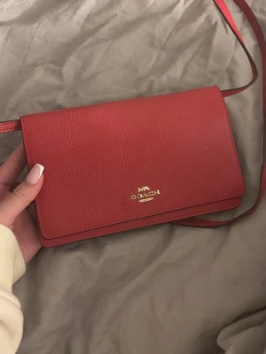 Röd axelväska från Coach - Snygg röd axelväska från Coach i  läder med guldfärgad logga framtill. Väskan har ett stilrent, rektangulärt format lock med magnetknäppning och justerbar axelrem. Invändigt finns dragkedjefack och flera fack för småsaker. Perfekt för dig som gillar klassisk men trendig stil. 