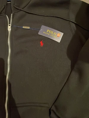 Svart hoodie från Polo Ralph Lauren - Säljer en svart hoodie från Polo Ralph Lauren med dragkedja och huva. Klassisk röd logga broderad på bröstet och två fickor framtill. Tillverkad i mjukt bomullsmaterial som känns skönt mot huden. Perfekt för en avslappnad och stilren look.