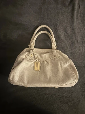 Beige handväska från Marc by Marc Jacobs - Säljer en snygg beige handväska från Marc by Marc Jacobs i mjukt, strukturerat läder. Väskan har dubbla handtag, guldfärgade metalldetaljer och en dekorativ tag framtill. Perfekt för dig som vill ha en stilren och rymlig väska.