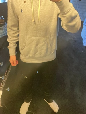 Grå hoodie från Polo Ralph Lauren - Snygg grå hoodie från Polo Ralph Lauren med klassisk känguruficka och dragsko i huvan. Liten broderad logga på bröstet. Tillverkad i mjukt bomullsmaterial som känns skönt mot huden. Perfekt för en avslappnad och stilren look.