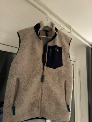 Patagonia fleeceväst - Säljer en beige fleeceväst från Patagonia med blå detaljer och dragkedjor. Västen har en hög krage, två sidofickor med dragkedja och en bröstficka i mörkblått tyg. Perfekt för lager-på-lager och riktigt skön att ha på sig. Kvitto ingår priset är inte hugget i sten