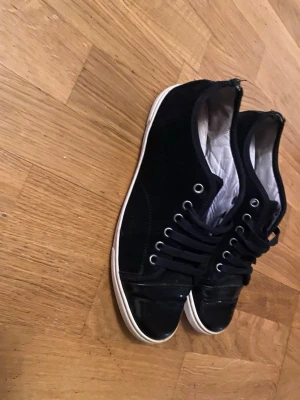Svarta sneakers från Lanvin - Snygga svarta sneakers från Lanvin med klassisk låg profil och vita sulor. Skorna har svarta skosnören, metallöljetter och ovandel i mocka. Insidan är fodrad i vitt tyg och designen är stilren och enkel, perfekt till streetwear eller casual outfits.