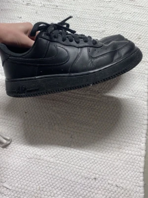 Nike Air Force 1 helsvarta sneakers - Klassiska Nike Air Force 1 sneakers i helsvart läder med svart sula och svarta skosnören. Ikonisk Nike Swoosh på sidan och AIR-detalj på hälen. Lågt skaft och perforeringar på tån för extra ventilation. Perfekta för en clean och stilren look.