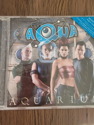 Aqua - Aquarius CD album - Säljer ett CD-album med titeln 'Aquarius' från popgruppen Aqua. Skivan kommer i ett genomskinligt plastfodral med färgglad framsida och låtlista på baksidan. Albumet innehåller 12 låtar, bland annat 'Cartoon Heroes' och 'Around The World'. Perfekt för dig som gillar 2000-talets popmusik.