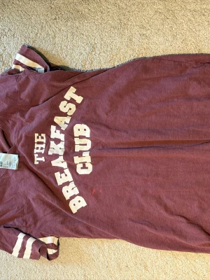 Vinröd pyjamaströja The Breakfast Club - Vinröd t-shirt i bomull med trycket 'The Breakfast Club' i vitt framtill. Kortärmad modell med vita ränder på ärmarna för en sportig vibe. Skön och avslappnad passform, perfekt till chill eller sovande.