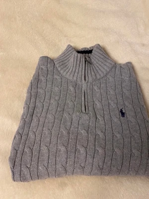 Grå kabelstickad tröja Polo Ralph Lauren - Kabelstickad grå tröja från Polo Ralph Lauren med halv dragkedja och hög krage. Tröjan har lång ärm, ribbade muddar och klassisk broderad logga på bröstet. Perfekt för lager-på-lager och snygg till jeans eller chinos.