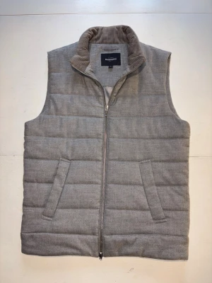 Grå dunväst från Dressmann - Snygg grå dunväst från Dressmann med quiltad design och dragkedja framtill. Västen har två snedställda fickor och en mjuk krage i manchestertyg. Perfekt för lager på lager och enkel att matcha med olika outfits. Storlek M. Skick 9/10.