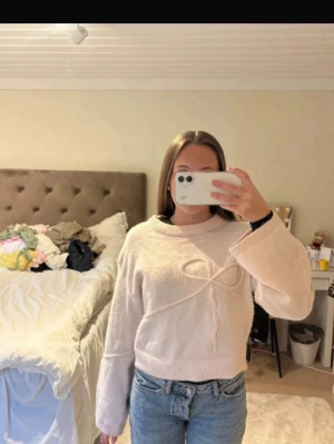 Ljusrosa stickad tröja med rosett - Supersöt ljusrosa stickad tröja med en dekorativ rosett framtill. Tröjan har rund halsringning, långa ärmar och ribbade muddar vid ärmslut och nederkant. Perfekt för dig som gillar en gullig och trendig stil.