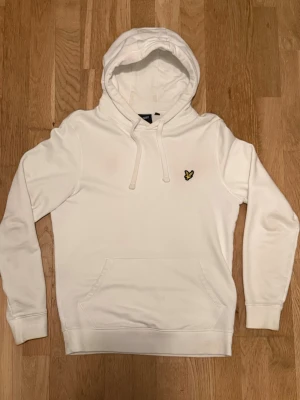 Vit hoodie från Lyle & Scott - Klassisk vit hoodie från Lyle & Scott med gul logga på bröstet. Hoodien har dragsko i huvan, magficka och ribbade muddar vid ärmar och nederkant. Perfekt för en clean och stilren look.