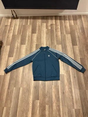 Blå Adidas track jacket - Säljer en blå track jacket från Adidas med klassiska vita ränder längs ärmarna och dragkedja framtill. Jackan har ståkrage, muddar vid ärmslut och nederkant samt Adidas-logga på bröstet. Perfekt för en sportig och avslappnad stil. Är storlek XS men jag har storlek S och den passar mig super bra.