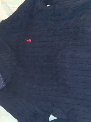 Mörkblå kabelstickad tröja Polo Ralph Lauren - Kabelstickad mörkblå tröja från Polo Ralph Lauren med rund halsringning och den klassiska röda logotypen broderad på bröstet. Tröjan har ribbade muddar och är perfekt för dig som gillar en tidlös och stilren look.