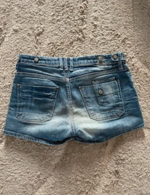 Blå denimshorts med fickor vintage - Säljer ett par klassiska blå jeansshorts i denim med två stora bakfickor och bälteshällor. Shortsen har låg midja och en snygg tvättad look med ljusare partier framtill och baktill. Perfekta till sommaren och lätta att matcha med olika toppar.
