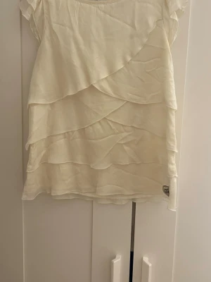 Vit volangblus från Pomp de Lux - Söt vit blus från Pomp de Lux med flera lager volanger framtill. Blusen har korta ärmar och rund halsringning. Storlek 140