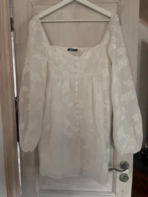 Vit blommig kortklänning med knappar - Söt vit kortklänning med blommigt mönster, långa puffärmar och knappar framtill. Klänningen har fyrkantig ringning och är tillverkad i ett lätt, transparent tyg med fodrad kropp. Perfekt för sommarens alla tillfällen och student😌 helt oanvänd!