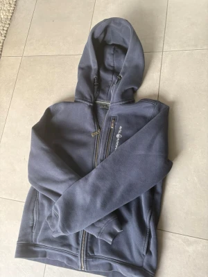 Mörkblå hoodie från Sail Racing - Mörkblå hoodie från Sail Racing med dragkedja framtill och två fickor. Klassisk huva och ribbade muddar vid ärmslut och nederkant. Tillverkad i mjukt och bekvämt material, perfekt för en avslappnad stil. Diskret logga på bröstet. Priset kan diskuteras vid snabb äffär 