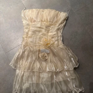 Klänning champagne färgad  - Superfin axelbandslös klänning i skimrande champagnefärg med volanger med en blomdetalj. Den är i krämvit/champagnefärgat tyg och är jättefin uppklädnad. Genomskinliga band följer med och pris kan diskuteras💕