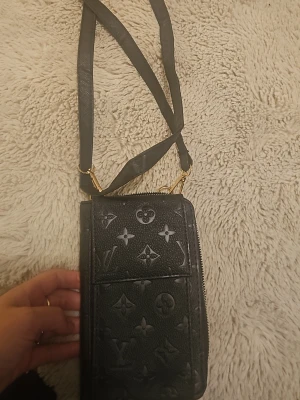 Svart plånbok med axelrem - Snygg svart plånbok med klassiskt monogrammönster i präglat läder. Plånboken har en praktisk axelrem, guldfärgade detaljer och flera fack, inklusive ett med dragkedja. Perfekt för dig som vill ha både stil och funktion.