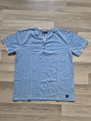 Ljusblå t-shirt från S.C.W. - Ljusblå t-shirt från S.C.W. med korta ärmar och knappar vid halsen. T-shirten har en avslappnad passform och är tillverkad i mjuk bomull. Detaljer som vit kant vid ärmslut och halsringning ger en snygg kontrast.