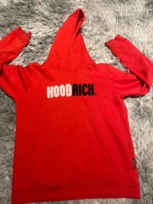 Hoodrich hoodie - Snygg röd hoodie från HOODRICH med stor vit och svart logga på bröstet. Klassisk känguruficka framtill och huva. Tillverkad i mjukt material som känns skönt mot huden. Perfekt för dig som gillar streetwear och vill sticka ut. Använd 4 gånger och ett tyg innanför tröjan som inte syns på utsidan har lossnat men man kan säkert göra något åt det.