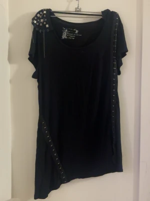Svart topp med nitar och detaljer - Cool svart topp med korta ärmar och asymmetrisk nederkant. Toppen har nitar och metallringar på ena axeln samt dekorativa detaljer längs sidan och framtill. Perfekt för dig som gillar en edgy stil.