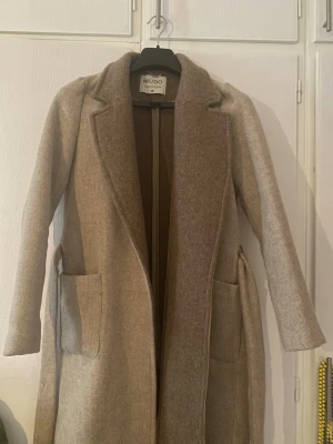 Beige ullkappa från Mudo - Stilren beige ullkappa från Mudo med klassisk krage, raka ärmar och två stora fickor framtill. Kappan har ett avtagbart bälte i midjan för en snygg siluett och är helfodrad. Perfekt för kyliga dagar när du vill vara både varm och trendig.
